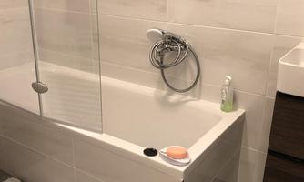 A modern bathroom with a bathtub and a glass shower partition. The walls are light tiled and there is a small rug in front of the tub.