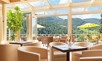 An elegant interior with large windows overlooking the mountains. Outside, there are sunny seating areas with umbrellas.