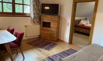 A cozy living space with a dining table and red chairs. In the background, a bedroom with a double bed can be seen.