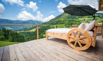 A cozy lounge chair on a wooden terrace overlooking the mountains. The sky is blue and decorated with white clouds.