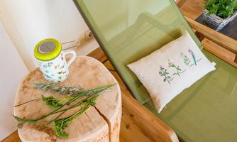 A cozy lounge chair is next to a wooden log with a coffee cup and fresh herbs. A decorative pillow with plant motifs completes the relaxing atmosphere.
