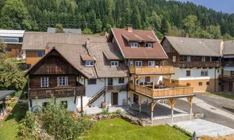 A charming wooden house with a balcony and a green meadow in front of it. Surrounded by wooded hills and other traditional buildings.