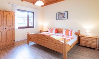 A bright bedroom with a double bed and wooden shelves. The walls are painted in soft colors, and the room has a large window with plenty of daylight.