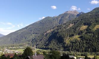 An impressive mountain landscape with wooded slopes and a clear blue sky. In the valley, small houses and meadows can be seen.
