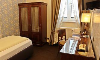 A cozy hotel room with a single bed, a desk, and a wardrobe. The room has bright curtains and decorative wallpaper.