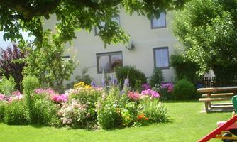 A beautiful garden with colorful flowers and lush greenery. In the background, there is a house and an outdoor seating area can be seen.