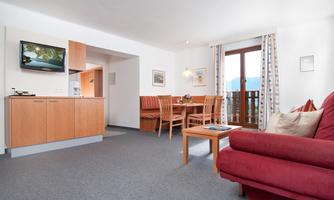 A bright living room with a red couch and a dining table. The kitchen is open and there is a balcony with a view.