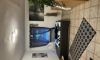 A cozy room with a large bed, plants, and simple furniture. Soft light falls through the window, and there are decorative pictures on the walls.