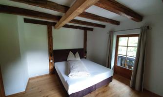 A simple bedroom with wooden flooring and exposed wooden beams. The bed has light bedding and a window provides daylight.