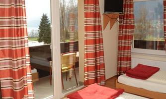 A bright room with two bed frames and red blankets. Large windows provide an outside view and plenty of daylight.