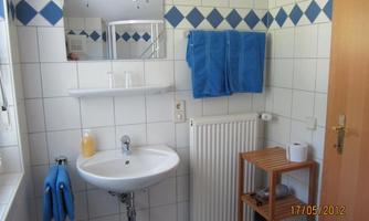 A modern bathroom with white tiles and blue accents. It features a sink, towels, and a wooden shelf.