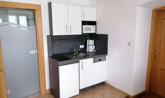 A modern kitchen unit with white cabinets, sink, and microwave. The floor is tiled and there is a door leading to another room.