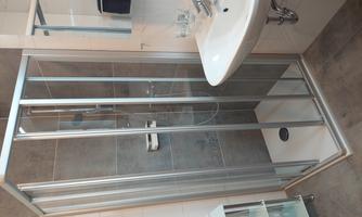 A modern bathroom with a glass shower cabin and a sink. The walls are light tiled, and there is a small shelf space.