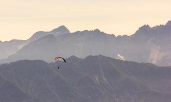 A paraglider soars over majestic mountains. The landscape is characterized by gentle hills and high peaks.
