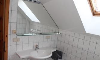 A modern bathroom with slanted walls, a sink, and a mirror. Bright tiles and a window provide natural lighting.