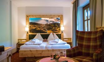 A cozy hotel room with two beds, a large window, and a view of a mountain landscape. The room is decorated with wood and an upholstered armchair.