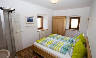 A bright bedroom with wooden flooring and a comfortable double bed. The walls are white, and there is a window as well as a wooden wardrobe.