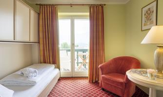 A cozy room with a single bed and a comfortable red armchair. Large windows provide a view outside and let in plenty of light.