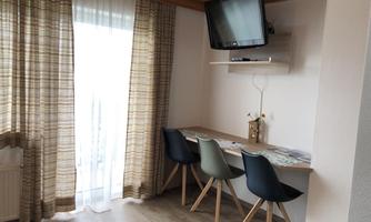 A cozy interior with a table and chairs. A television hangs on the wall and large windows let in plenty of light.