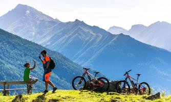 Two cyclists are standing in the mountains while taking a break. In the background, impressive mountains and their bikes can be seen.