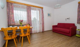 A bright living space with a red sofa and a dining table. Light curtains hang in front of the windows, and fresh flowers adorn the table.