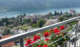 A picturesque view of a lake surrounded by mountains, with colorful flowers on a balcony in the foreground. The landscape features small houses and a clear blue sky.