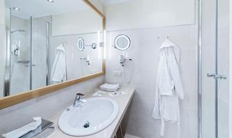 A modern bathroom with a large mirror, sink, and a shower. White bathrobes hang on the wall, and the decor is bright and inviting.