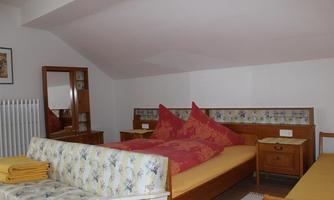 A simple bedroom with a comfortable bed and two cushions in red colors. There is a seating area and a wardrobe with a mirror.