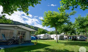 A campsite with various caravans and tents in a green environment. Trees provide shade and the clouds in the blue sky are visible.