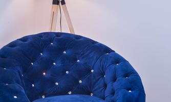A round, blue armchair with a velvety surface stands next to a floor lamp. The lamp has a beige shade and provides cozy light in the room.
