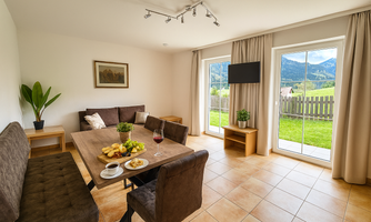 A cozy living area with a dining table and a seating area. Large windows provide a view of the green landscape and the mountains.