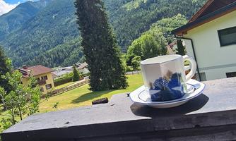 A cup of coffee on a table with a view of green mountains and a blue sky. In the background, trees and houses can be seen.