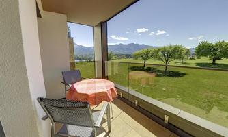 A cozy balcony with a table and chairs. In the background, green meadows and mountains can be seen under a clear blue sky.