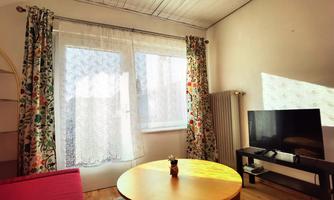 A bright living room with colorful curtains and large windows. In the room, there is a round table and a television.