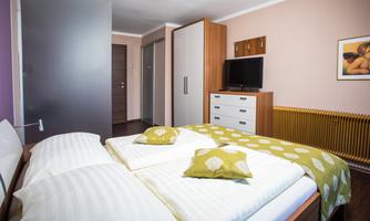 A cozy bedroom with a double bed, two pillows, and a decorative blanket. There is a television on the wall, and there are cabinets as well as a radiator.