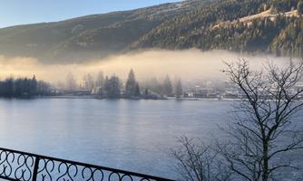 A tranquil winter landscape with a frozen lake and gentle mist. In the background, forested mountains rise under a clear blue sky.