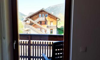 A window view of a wooden house in the mountains. In the foreground, there is a chair on the balcony.