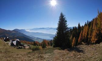 A picturesque mountain landscape with radiant sunshine. In the foreground are some wooden houses and trees in autumn colors.