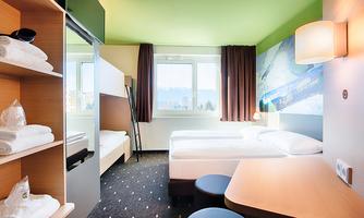 A modern hotel room with two beds and a large window. The walls are painted green, and there is a small table as well as a shelf with towels.