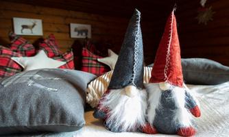 Two cute gnomes with long hats are sitting on a white pillow. In the background, cozy pillows and wall pictures can be seen.