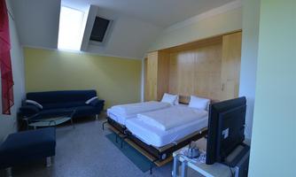 A simple room with two single beds, a sofa, and a television. The room has a light wall color and large window light.