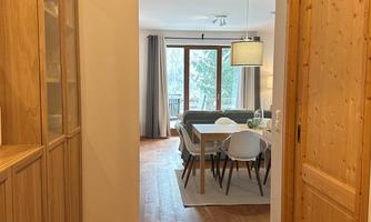 A bright hallway with wooden paneling leads to a cozy living area. Large windows let in light and provide a view of nature.