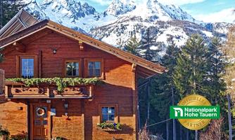 A cozy wooden house in front of majestic, snow-covered mountains. Surrounded by dense coniferous trees and a clear sky.