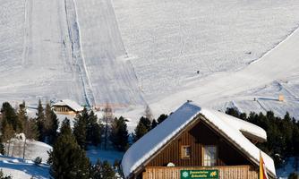A cozy wooden house in the snow, surrounded by a wintry landscape. In the background, a snow-covered slope extends.