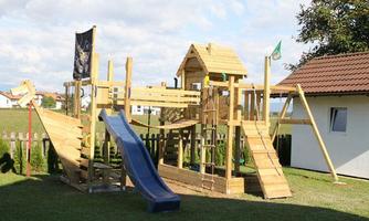 A large wooden playground with slides and climbing elements. In the background, there is a house and a green meadow.