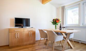 A cozy dining area with a wooden table and chairs. Next to the table is a vase with flowers and a bottle of wine.