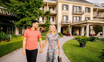 A happy couple strolls hand in hand in front of an elegant building. The surroundings are green and inviting.