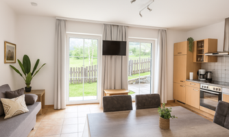 A bright, inviting apartment with a large dining table and a modern kitchen. Large windows provide a view of the green landscape outside.