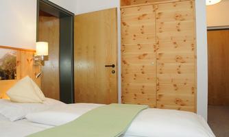 A cozy bedroom with a soft bed and a green blanket. The walls are light and there is a wardrobe made of light wood.