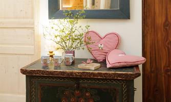 A decorative table with a pretty flower vase and two cups. Next to it lies a red heart-shaped pillow and a small wooden board.